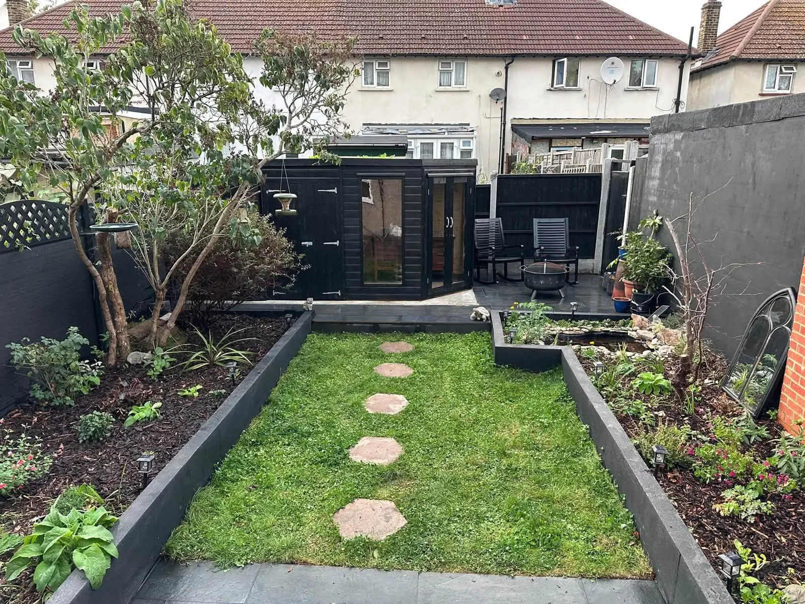 A finished back garden in London: narrow lawn with hexagonal stepping stones, black sleeper beds and a black garden room